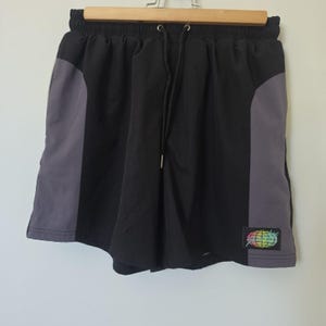 May include: Black athletic shorts featuring dark gray side panels and an elastic waistband with a black drawstring. A small rectangular patch on the lower right leg displays a colorful graphic of a globe with a lightning bolt design.