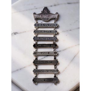 Puede incluir: Una insignia de metal plateado con un escudo superior con las letras griegas ΦΔΘ, seguida de una cadena vertical de ocho placas rectangulares grabadas con nombres de ciudades y años: Dallas 1988, Kansas City 1990, Arlington 1992, Scottsdale 1994, Orlando 1996, Marina Del Rey 2004, Kansas City 2006 y Paradise Valley 2008.
