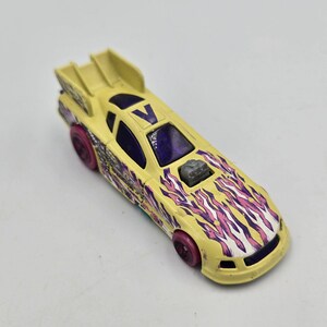 May include: A yellow die-cast toy race car featuring a large rear spoiler, purple tinted windows, and a purple letter V on the roof. The hood and sides are decorated with a purple and pink flame pattern, and the car has four small pink wheels.