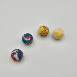 May include: Three spherical bouncy balls with smooth surfaces. One ball features a marbled pattern of blue, red, and white. Another ball displays a marbled mix of yellow, orange, and black. The third ball is a solid cream color with yellow swirls.