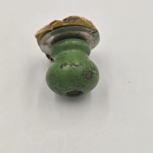 Peut inclure: Un petit bouton arrondi de couleur vert clair avec une finition patinée et des taches sombres. Le bouton présente une plaque de base en métal argenté et un support adhésif circulaire de couleur beige fixé sur le dessus, montrant des signes d'usure.