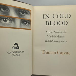May include: The title page of the book In Cold Blood by Truman Capote, published by Random House New York. The page features a black and white graphic of two pairs of eyes in a rectangular frame, with the book title and author name in black text.