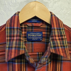 Op de afbeelding: Een overhemd met lange mouwen en knoopsluiting in een rood, blauw, geel en zwart ruitpatroon. Het overhemd heeft een puntige kraag en een blauw Pendleton-merklabel in de nek, wat aangeeft dat het is vervaardigd door Pendleton Woolen Mills.