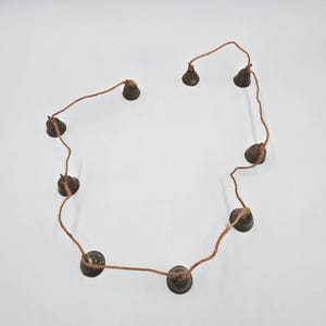 May include: A string of eight small, dark, metallic bells attached to a thin, light brown twine. The bells are spaced out along the cord, each featuring a simple, conical shape with a dark, weathered finish, arranged in an irregular loop.