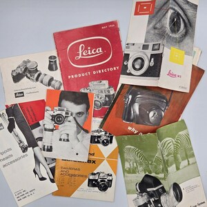 Puede incluir: Una colección de folletos y directorios de productos Leica antiguos con fotografías en blanco y negro de cámaras, lentes y accesorios. Los materiales impresos incluyen portadas rojas, naranjas y verdes con texto como Leica Product Directory.