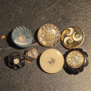 Könnte beinhalten: Eine Sammlung von sieben verschiedenen runden Knöpfen mit unterschiedlichen Designs, darunter ein blauer Wirbelknopf, ein transparenter Glitzerknopf, ein Knopf mit weißen Halbmondformen, ein schwarzer Filigranknopf, eine kleine transparente Perle, ein cremefarbener Knopf mit einem zentralen Kristall und ein schwarzer Knopf mit einem strukturierten Kristallzentrum.