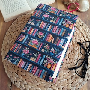 Könnte beinhalten: Ein Stoff-Buchumschlag mit dunkelblauem Hintergrund und einem sich wiederholenden Muster aus Bücherregalen, gefüllt mit bunten Büchern und Blumenarrangements in Vasen. Der Umschlag ist rechteckig und enthält ein schmales, rosa Lesezeichenband.