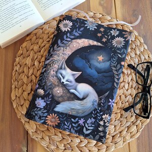 May include: A black book cover featuring a white fox asleep on a crescent moon. The moon is surrounded by stars and clouds. The background is a dark blue with white flowers.