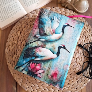 Könnte beinhalten: Ein Buchumschlag mit einem Aquarellgemälde von zwei Kranichen mit roten Kronen und langen schwarzen Schnäbeln, vor einem blau-rosa Hintergrund mit Lotusblumen. Ein rosa Leseband ist befestigt. Eine schwarze Brille liegt daneben.