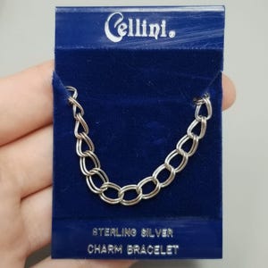 May include: A silver-toned chain link bracelet displayed on a dark blue velvet card. The card features the white text Cellini at the top and the text STERLING SILVER CHARM BRACELET at the bottom. The bracelet consists of oval-shaped metal links.