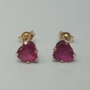 Puede incluir: Un par de pendientes de botón con gemas de color rosa intenso en forma de corazón montadas en metal dorado. Cada pendiente tiene un engaste de garras, un poste dorado y un cierre de mariposa visible en la parte posterior.