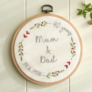 May include: A circular wooden embroidery hoop containing white fabric with grey embroidered text reading 40 years Mum & Dad and the date 14.6.1986. The text is surrounded by a delicate embroidered wreath of green leaves and small red berries.