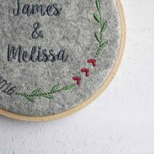 Puede incluir: Un c&iacute;rculo de fieltro gris con los nombres "James & Melissa" bordados en hilo negro. Una vid verde frondosa con bayas rojas est&aacute; bordada alrededor del borde.