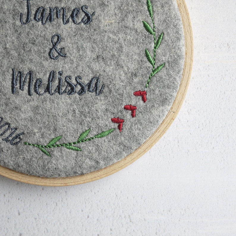 Puede incluir: Un c&iacute;rculo de fieltro gris con los nombres "James & Melissa" bordados en hilo negro. Una vid verde frondosa con bayas rojas est&aacute; bordada alrededor del borde.