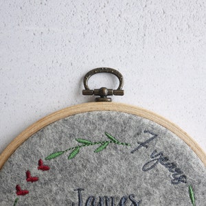 Puede incluir: Un aro de bordado de fieltro gris con un marco de madera y un anillo de lat&oacute;n. El aro presenta un dise&ntilde;o de hojas verdes, corazones rojos y el texto "1 a&ntilde;o" y "James".