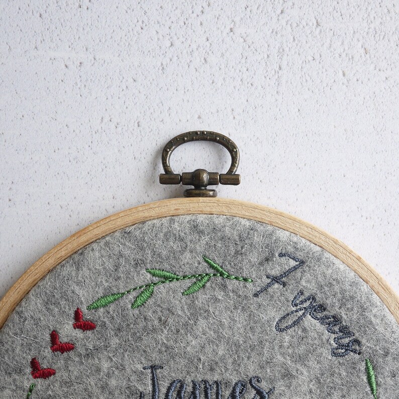 Puede incluir: Un aro de bordado de fieltro gris con un marco de madera y un anillo de lat&oacute;n. El aro presenta un dise&ntilde;o de hojas verdes, corazones rojos y el texto "1 a&ntilde;o" y "James".