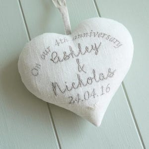 Puede incluir: Un adorno de tela en forma de corazón de color blanco roto con texto bordado en gris que dice: On our 4th anniversary Ashley & Nicholas 24.04.16. El adorno incluye un lazo de tela en la parte superior para colgarlo y tiene una textura tejida.