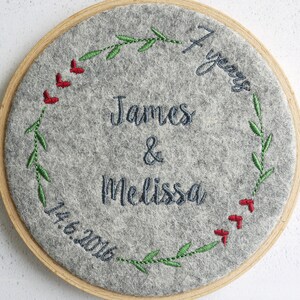 Puede incluir: Un c&iacute;rculo de fieltro gris con un marco de madera, bordado con una corona de hojas verdes y bayas rojas. El texto "7 a&ntilde;os" est&aacute; bordado en la esquina superior derecha. El texto "James & Melissa" est&aacute; bordado en el centro. La fecha "14.6.2016" est&aacute; bordada en la esquina inferior izquierda.
