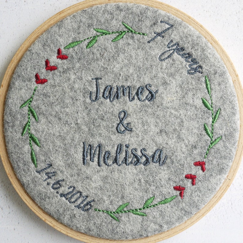 Puede incluir: Un c&iacute;rculo de fieltro gris con un marco de madera, bordado con una corona de hojas verdes y bayas rojas. El texto "7 a&ntilde;os" est&aacute; bordado en la esquina superior derecha. El texto "James & Melissa" est&aacute; bordado en el centro. La fecha "14.6.2016" est&aacute; bordada en la esquina inferior izquierda.