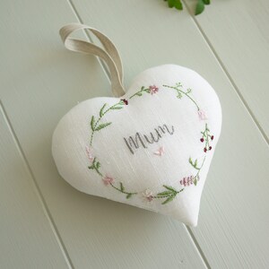 Puede incluir: Almohada blanca en forma de corazón con una corona de flores bordada alrededor del borde. La corona está hecha de hojas verdes y flores rosas. La palabra "Mamá" está bordada en el centro de la almohada con hilo gris.