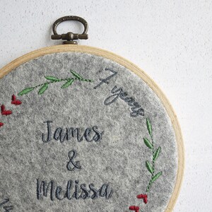 Puede incluir: Aro de fieltro gris bordado con un marco de madera y un anillo de lat&oacute;n. El aro presenta un dise&ntilde;o floral con bayas rojas y hojas verdes. El texto "James & Melissa 4 a&ntilde;os" est&aacute; bordado en hilo negro.