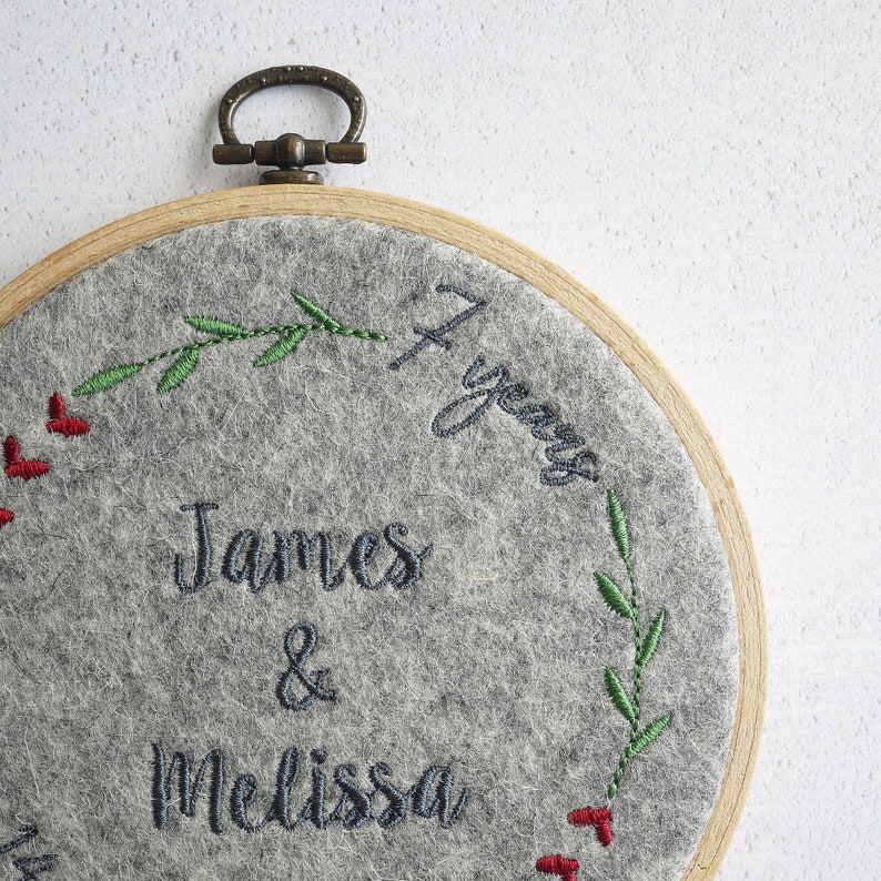 Puede incluir: Aro de fieltro gris bordado con un marco de madera y un anillo de lat&oacute;n. El aro presenta un dise&ntilde;o floral con bayas rojas y hojas verdes. El texto "James & Melissa 4 a&ntilde;os" est&aacute; bordado en hilo negro.