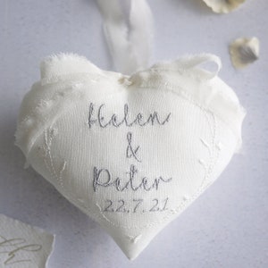 Puede incluir: Un corazón de tela blanco con bordes deshilachados y una cinta blanca. El centro está bordado con el texto gris "Helen & Peter" y la fecha "22.7.21" en letra cursiva, rodeado por un delicado patrón bordado de ramas con hojas.