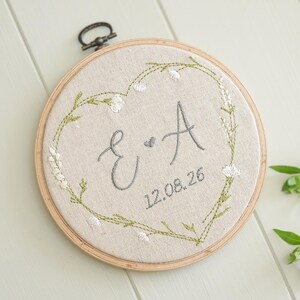 May include: A circular embroidery hoop with linen fabric, displaying the letters "E & A" and the date "12.08.26" in grey thread. The letters are within a heart-shaped design of green leaves and white flowers. The hoop is made of wood.