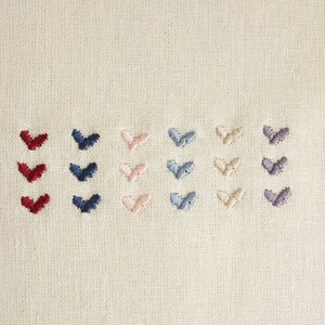 Puede incluir: Primer plano de una tela blanca con filas de corazones bordados en diferentes colores: rojo, azul marino, rosa claro, azul claro, beige y gris.
