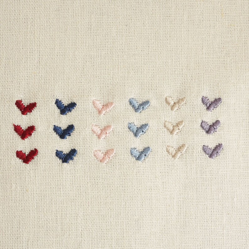 Puede incluir: Primer plano de una tela blanca con filas de corazones bordados en diferentes colores: rojo, azul marino, rosa claro, azul claro, beige y gris.