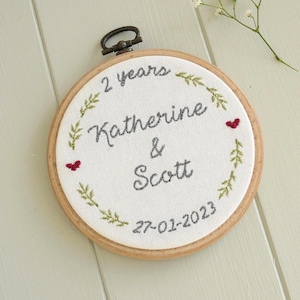 Puede incluir: Un bordado circular con los nombres "Katherine & Scott", la fecha "27-01-2023" y la frase "2 years". El diseño incluye hojas verdes, corazones rojos y un aro de madera con un colgador de metal.