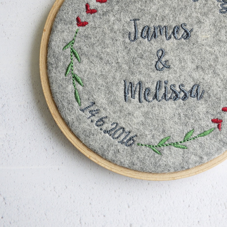 Puede incluir: Arte de bordado en aro con fondo de fieltro gris, con una corona de hojas verdes y corazones rojos. El texto "James & Melissa 14.6.2016" est&aacute; bordado en hilo azul.