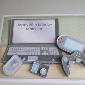 May include: A birthday card with a laptop, handheld game console, game controller, camera, and mobile phone illustrations. The laptop screen displays "Happy 80th Birthday Malcolm". The background is light green with white polka dots. A unique birthday card.