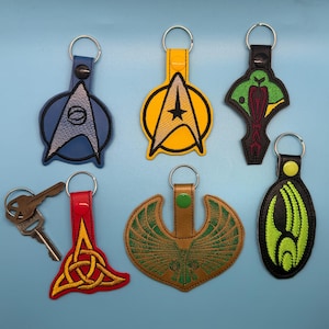May include: A collection of embroidered keychains in various shapes and colors. Designs include a blue Starfleet insignia, a yellow Star Trek emblem, a green and red design, a red and gold Celtic knot, a brown and green winged design, and a black and green design.