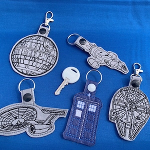 May include: Five different keychains featuring popular science fiction characters and spaceships. The keychains are made of gray fabric with black embroidery. The characters include the Death Star, the Millennium Falcon, the TARDIS, the Starship Enterprise, and a Serenity spaceship.