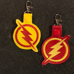 May include: Two keychains with the Flash logo. One keychain is yellow with a red lightning bolt and the other is red with a yellow lightning bolt.