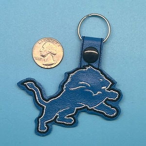 Puede incluir: Un llavero azul y plateado con forma de león con un llavero plateado. El llavero está hecho de un material texturizado y presenta un contorno negro. Una moneda de veinticinco centavos estadounidense está al lado del llavero para comparar el tamaño.