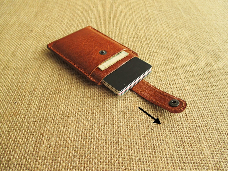 K&ouml;nnte beinhalten: Eine braune Ledergeldb&ouml;rse mit einer Klappe, die mit einem Druckknopf geschlossen wird. Die Geldb&ouml;rse hat einen Riemen, der zum Befestigen an einem G&uuml;rtel oder einer Tasche verwendet werden kann.