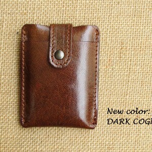 K&ouml;nnte beinhalten: Eine braune Ledergeldb&ouml;rse mit einem Klappverschluss und einem Metallknopf. Die Geldb&ouml;rse befindet sich auf einem braunen Jutesack-Hintergrund. Neue Farbe: DARK COGNAC