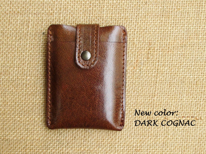 K&ouml;nnte beinhalten: Eine braune Ledergeldb&ouml;rse mit einem Klappverschluss und einem Metallknopf. Die Geldb&ouml;rse befindet sich auf einem braunen Jutesack-Hintergrund. Neue Farbe: DARK COGNAC