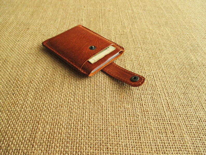 K&ouml;nnte beinhalten: Eine braune Ledergeldb&ouml;rse mit einem Klappverschluss und einem Druckknopf. Die Geldb&ouml;rse ist ge&ouml;ffnet und eine Kreditkarte ist im Inneren sichtbar.