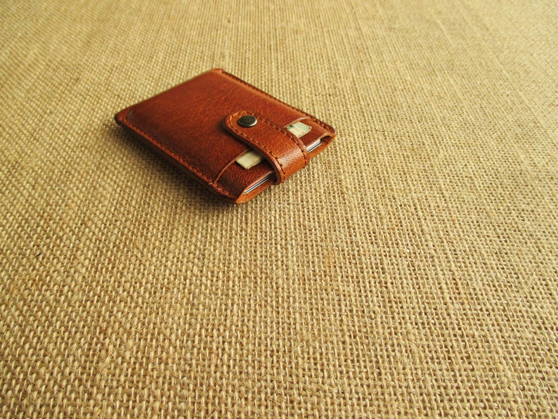 K&ouml;nnte beinhalten: Eine braune Ledergeldb&ouml;rse mit einem Klappverschluss und einem Metallknopf. Die Geldb&ouml;rse liegt auf einer braunen Jutestoffoberfl&auml;che.