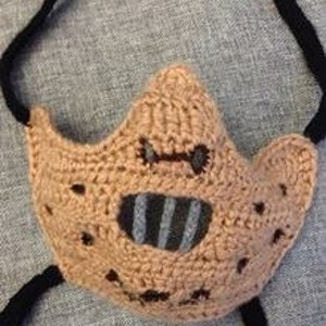 Op de afbeelding: Gehaakte gezichtsmasker met een bruin en zwart ontwerp. Het masker heeft een mondopening met een gestreept patroon en gaten rond de ogen.