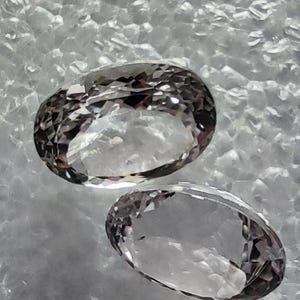 May include: Two clear, faceted, oval-shaped gemstones with a slight purple hue. The gemstones are on a white, textured surface.