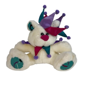May include: A plush white teddy bear wearing a jester hat with teal, purple, and magenta accents. The bear has a magenta nose, black eyes, and teal accents on its ears and feet. The bear is in a sitting position.