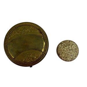 Peut inclure: Deux poudriers anciens en laiton. Le plus grand est rond avec des motifs floraux et géométriques. Le plus petit compact a un motif floral sur le couvercle. Les deux sont posés sur un fond blanc.