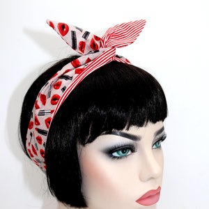 May include: A red and white headband with a lipstick print. The headband is tied in a bow on top of the head. The headband is worn by a mannequin with black hair.