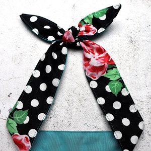 May include: A black and white polka dot headband with a teal lining. The headband features a floral pattern with red roses and green leaves. The headband is tied at the top with two long, pointed ends.