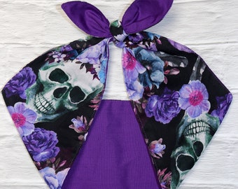 May include: A purple and black headband with a floral and skull print. The headband features a knot at the top with two purple bow-like extensions. The main body of the headband has a black background with purple flowers and skull graphics.