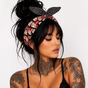 May include: A fabric headband featuring a repeating cartoon character print with black hair and red clothing on a beige background, topped with a black and white polka dot bow tie detail. The headband is worn across the forehead.